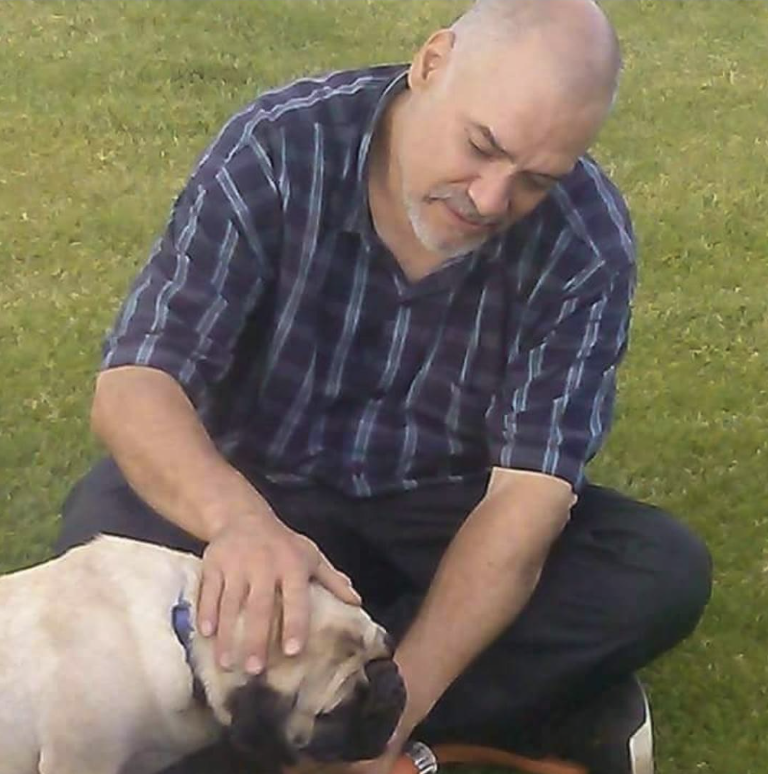 A middle-aged man in a plaid shirt is sitting on the grass, gently petting a pug. He has a bald head and is looking affectionately at the dog. The setting appears to be a park with green grass, suggesting a relaxed and joyful interaction between the man and the animal.